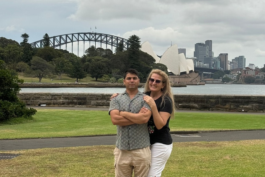A couple posing in a park with the Sydney Harbour Bridge and Opera House behind them. 