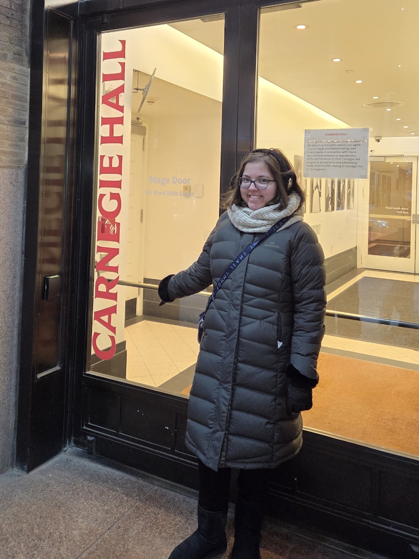 Stefania, wrapped up in a long puffer coat, stands in front of a door pointing at a sign saying Carnegie Hall