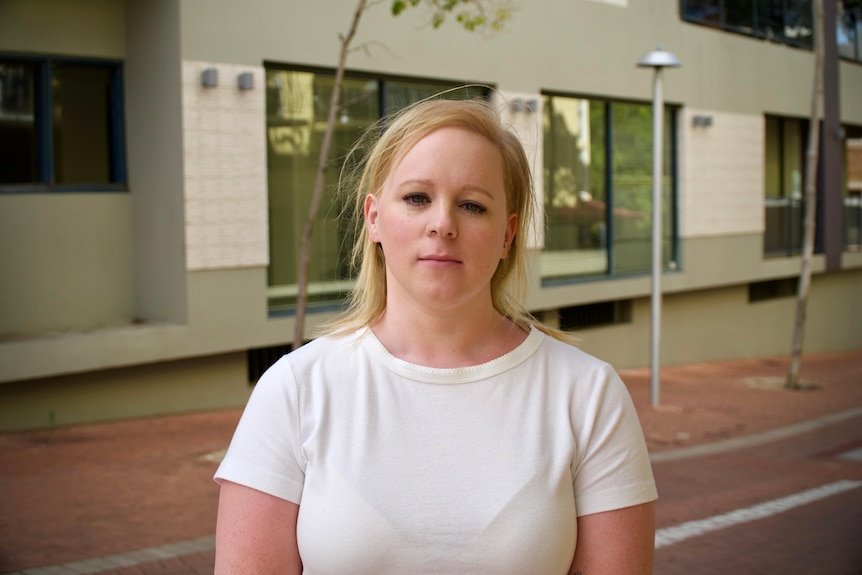 A young blonde woman stands outside a larhge building in a city.