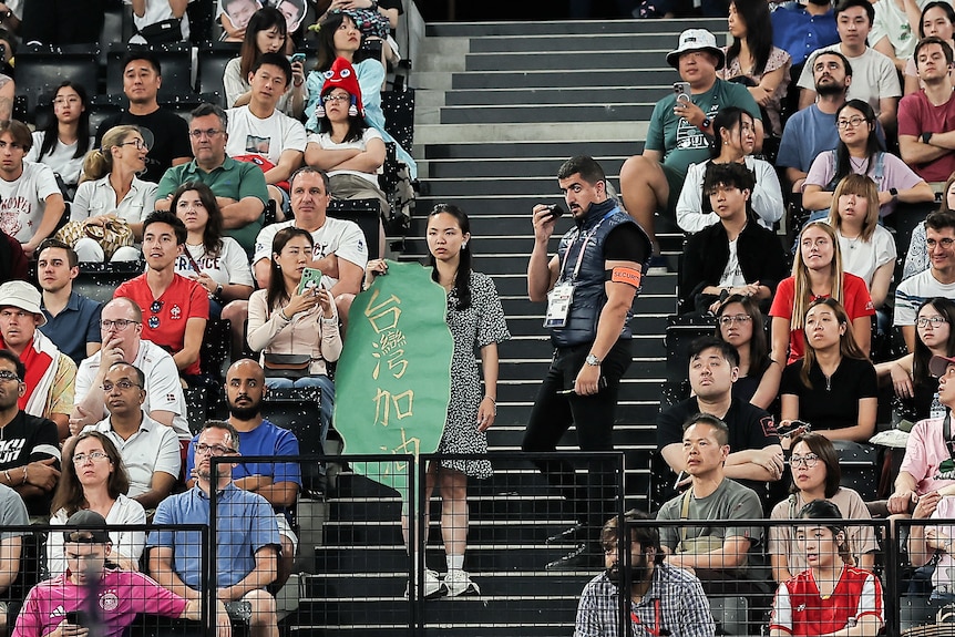 A woman holding a protest sign stands in a large crowd inside a stadium