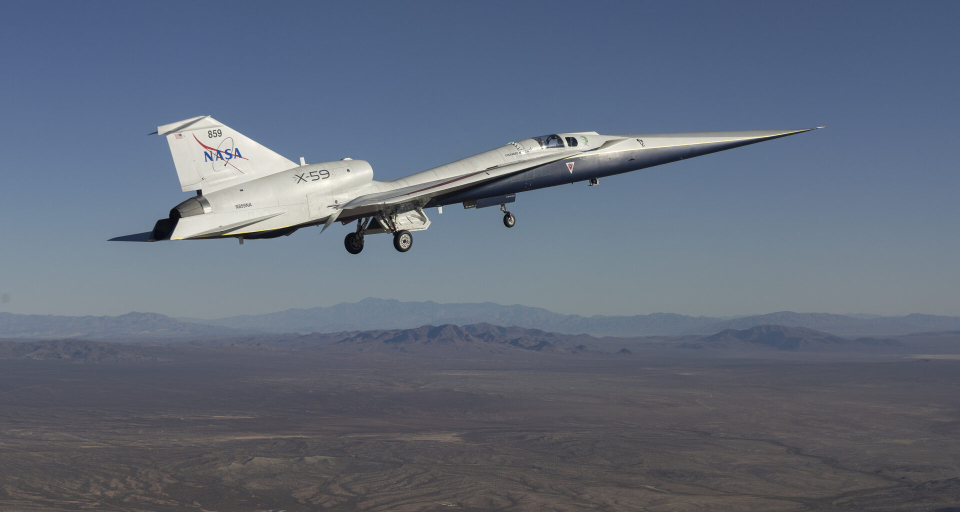 Image of a sleek, white airplane with a sharp, pointed nose flies above arid mountains. The plane's wheels are down. NASA is painted in blue lettering on its tail. The X-59 has a role in NASA's Flight Demonstrations and Capabilities (FDC) project.
