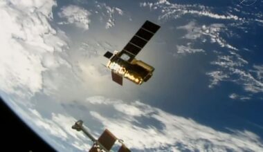 a cylindrical golden spacecraft with a pair of long, rectangular solar arrays extended is seen from above with earth in the background