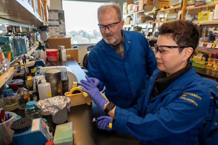 Two researchers in blue lab coats, gloves and protective glasses at a laboratory bench amid shelves of scientific supplies.