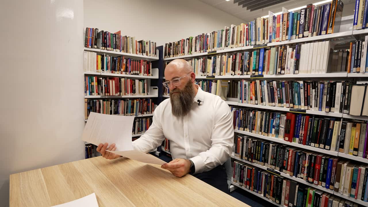 A man wearing a white shirt sits at a desk in a public library and reads from a document he is holding. There are shelves of books close behind him.