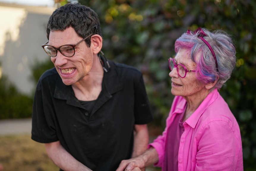 Mature woman in all-pink outfit and pink thick-rimmed glasses looks at her adult son, who also wears glasses, as he smiles big