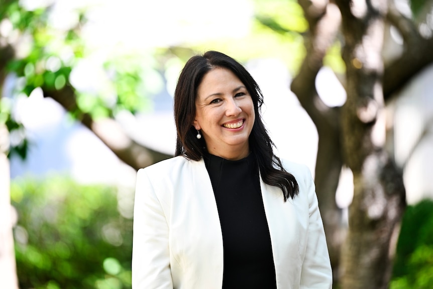 A woman smiles in a white blazer in front of a large tree