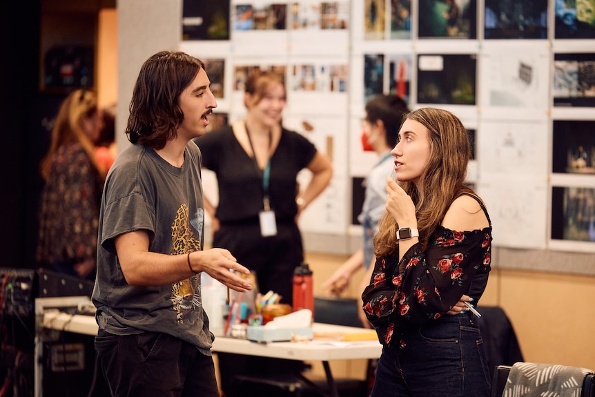 A young man with long hair, a moustache and a t-shirt talking to a young woman with long hair and an off-the-shoulder top.