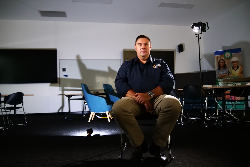 A man sitting on a chair in a room with a whiteboard, chairs and mounted light.