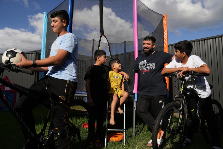 Man stands in a backyard next to a trampoline. Four boys of various ages next to him standing and on bikes