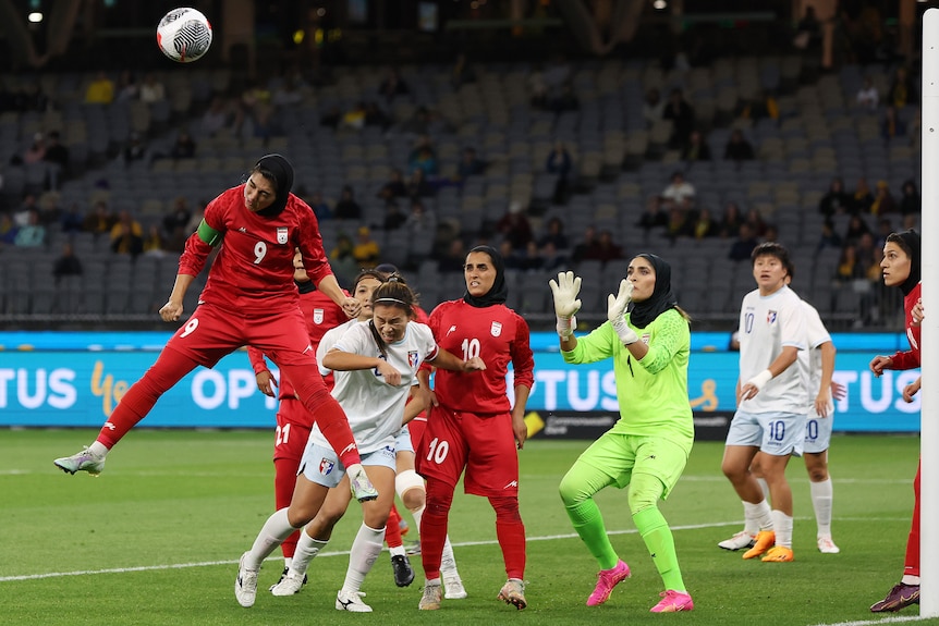A member of the Iran women's football team is in mid air after heading the ball, she's surrounded by other players.