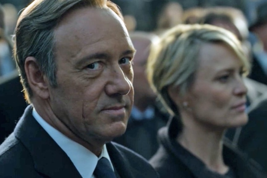 White man in business suit glances at the camera guiltily. An attractive woman with cropped hair is in the background
