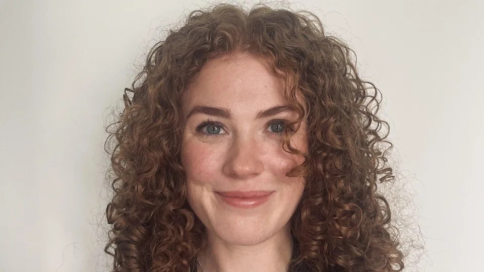 Gemma is standing in front of a white background and is smiling into the camera. She is a young woman with curly red hair. She has blue eyes.