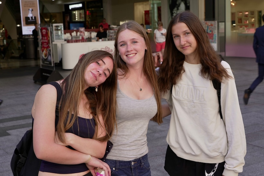 Three teenage girls stand together in a shopping area, smiling.