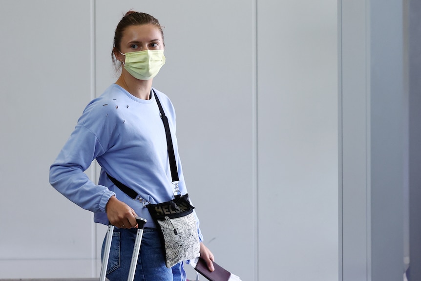 A woman looks sternly while wearing a facemask and carrying luggage at an airport