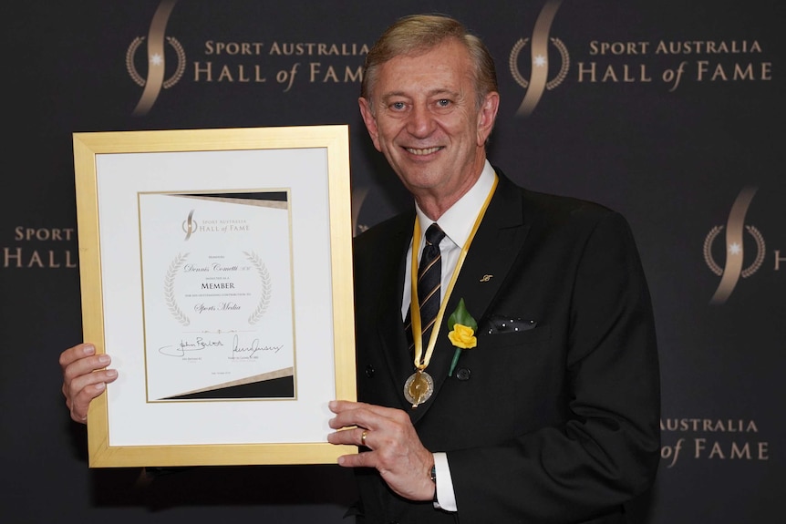 A smiling, middle-aged man in a dark suit holds up a framed certificate.