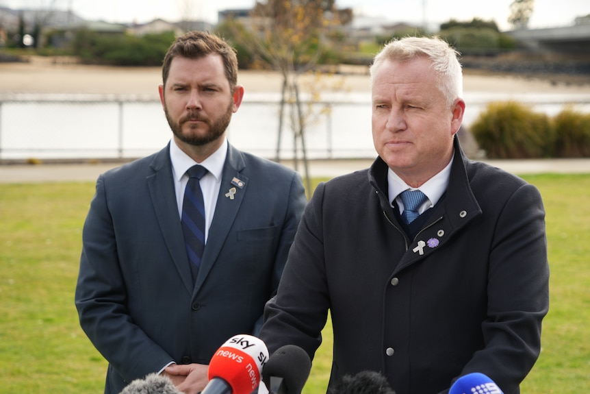 Two men in suits stand on a football field in front of microphones, addressing the media.