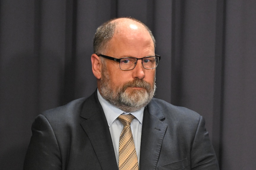 A man in a suit and glasses with grey beard looks sternly at camera.
