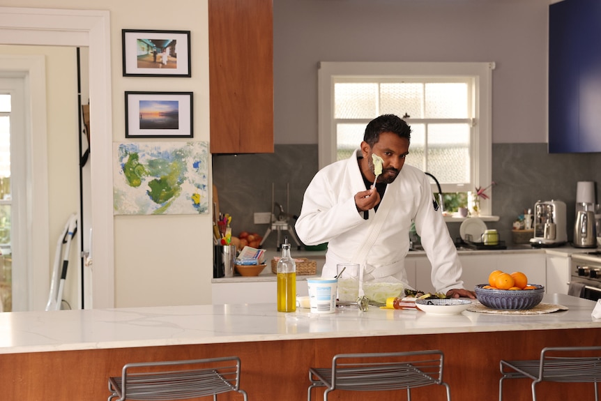 Nazeem Hussain stands in a home kitchen in a robe, wiping yoghurt onto his face.