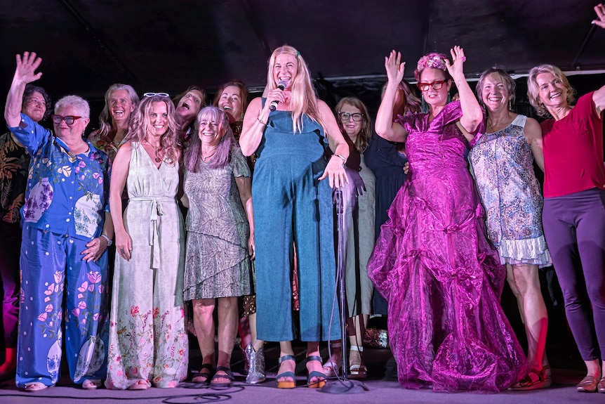 A group of women stand on stage smiling and waving.