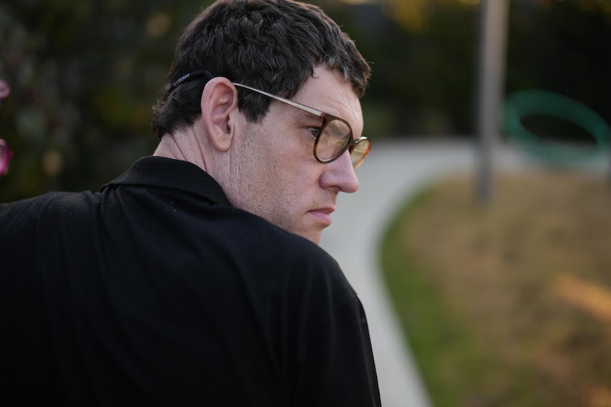 45yo man with black T-shirt and thick-rimmed glasses, outside setting, looks over his shoulder