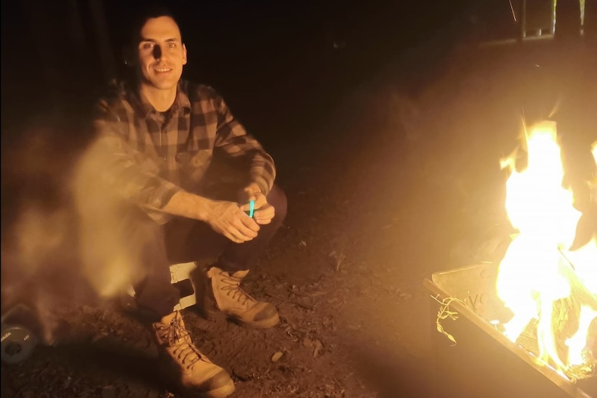 A man sits by a fire looking straight at the camera. 