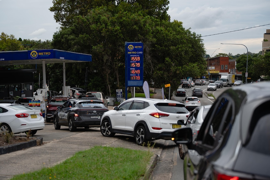 Cars file into a service station, green trees behind, cloudy evening sky.