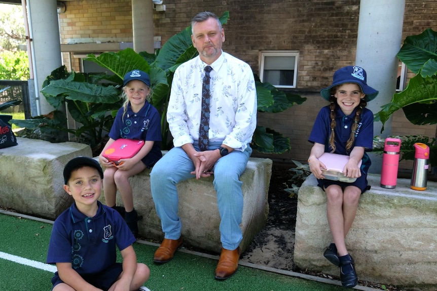 A man dressed in a white shirt and blue tie sitting on a sandstone block with two girls either side and a boy sitting in front.