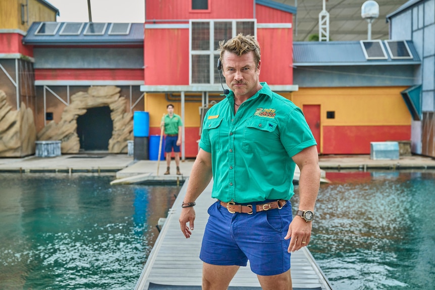 A TV still of Luke Hemsworth, 45, dressed as a celebrity croc wrangler, standing proudly beside an aquarium pool.