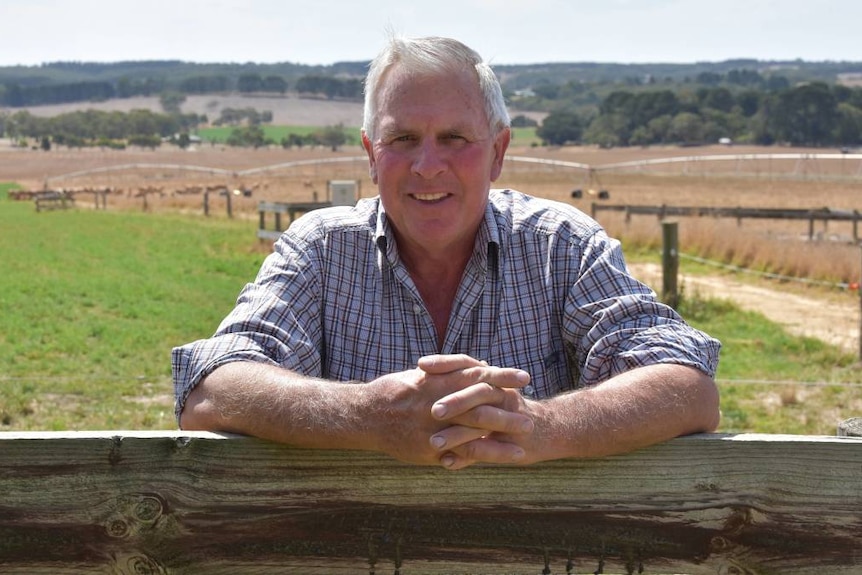 Robert, a middle-aged white-haired fair-skinned man, smiles as he leans on a farm fence post with his hands together 