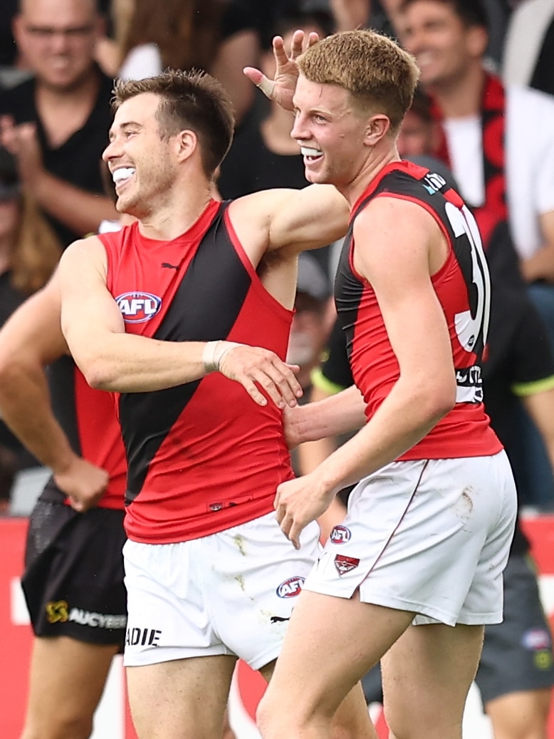 A young Essendon AFL player smiles in celebration after kicking a goal as a veteran player smiles and pats him on the head.