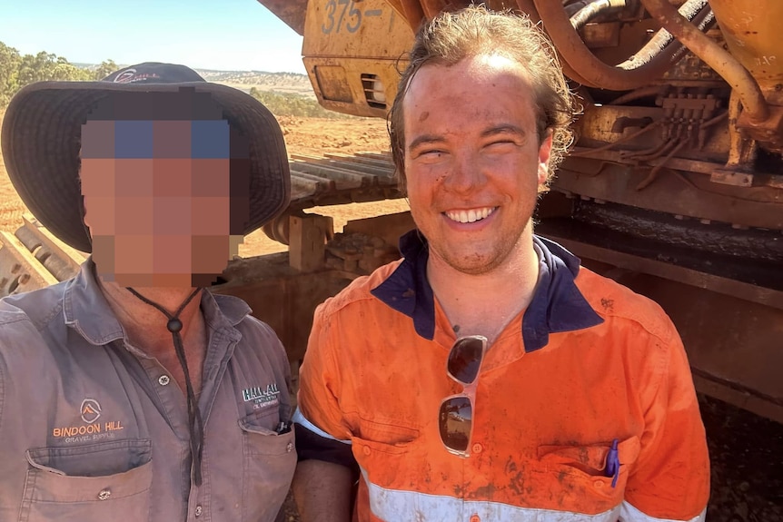 Two men smile at the camera, one in high-vis, with a digger behind them