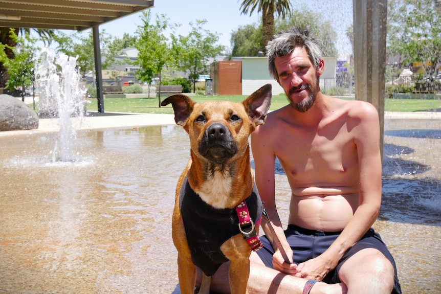 A shirtless man and his dog sit in front of a fountain.