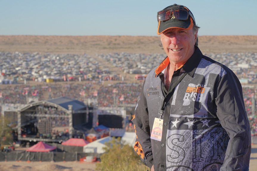 A man in a cap stands on a dune with a big festival crowd behind him