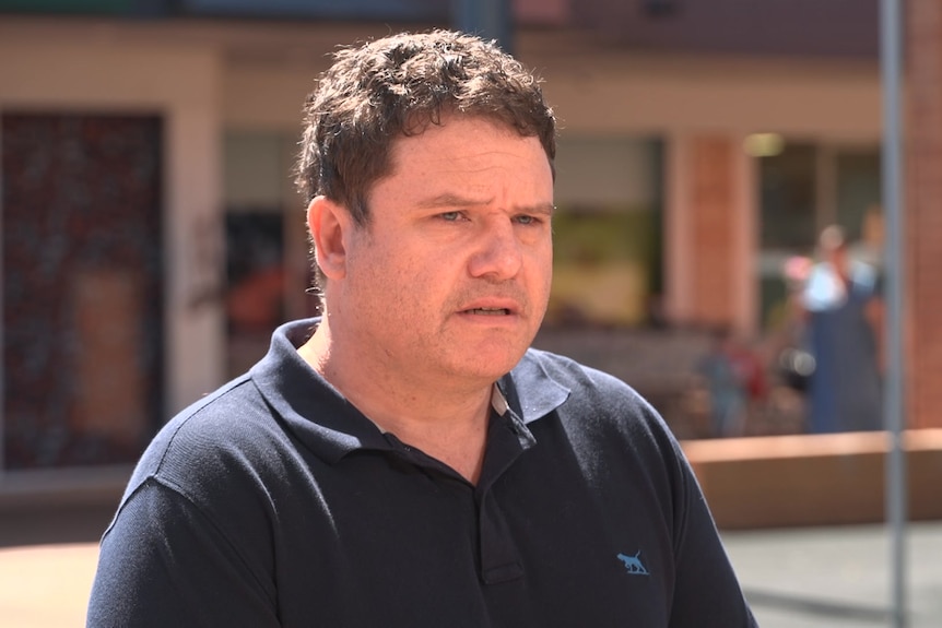 A man with short brown hair stands outdoors looking serious.