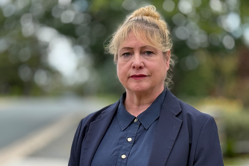 A woman with blonde hair tied back stands outside, looking into the camera, wearing a blue collared blouse and blue blazer.