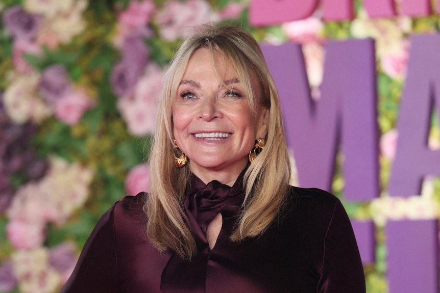 Helen Fielding wears a burgundy satin evening dress and poses with one hand on her hip on a red carpet.