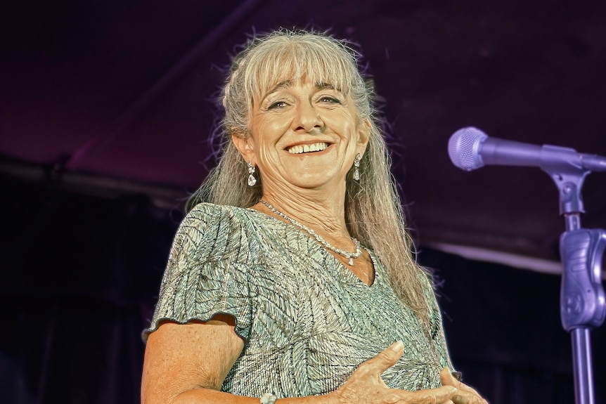 A  smiling woman in a glittering dress stands in front of a microphone.