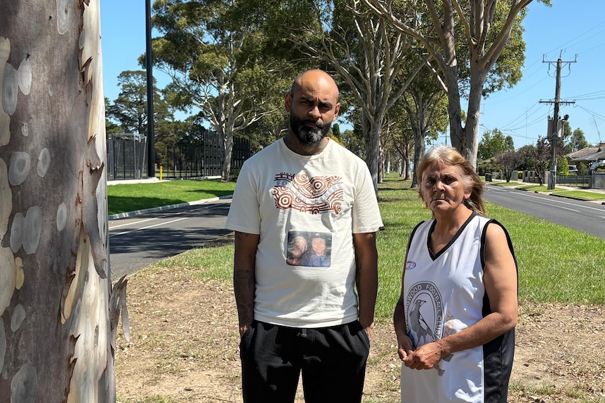 An Indigenous man and his mother stand on a median strip in a regional town looking pensive.