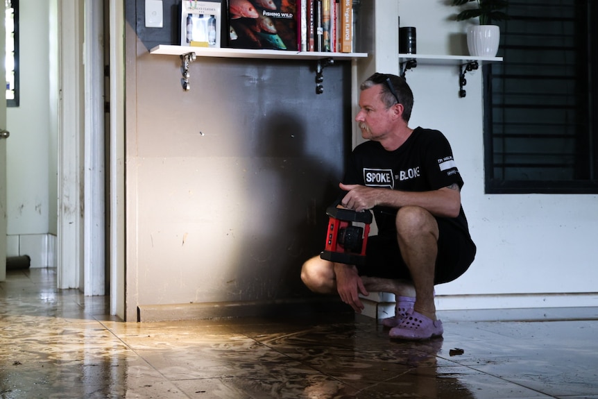 A man kneels and shines a torch across the floor of a room that is covered in mud