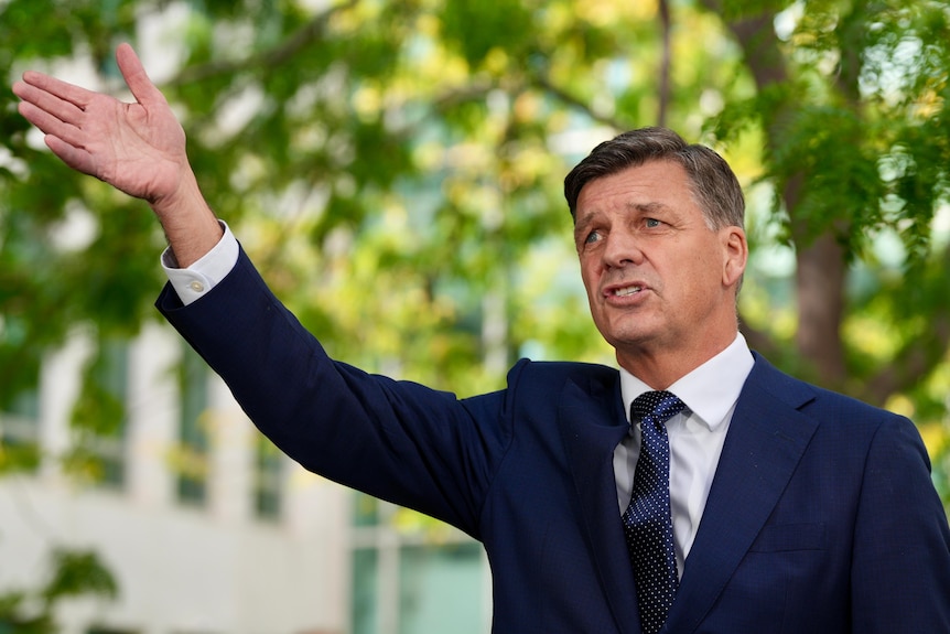 A man in a suit extends his hand in exclamation while speaking to a group of journalists.