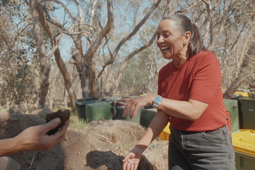 Myf points at Hamish's hand holding compost out to her as they stand in a field packed with bins.