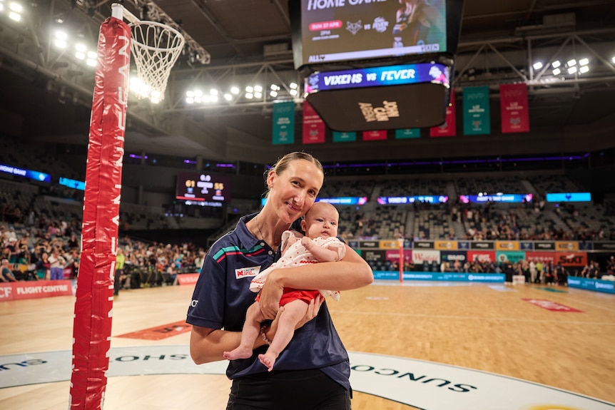 A player stands on court in front of a netball hoop and holds her baby daughter in her arms 