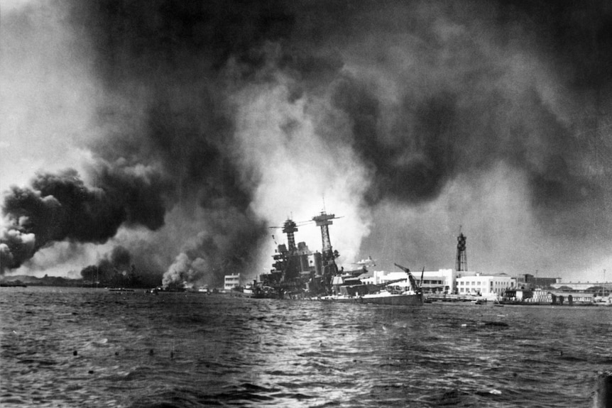 A black and white photo showing a burning ship sinking into a harbour.