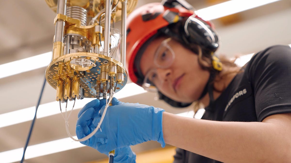 an engineer in blue gloves and safety gear screws a cable into the bottom of a gold-plated dilution refrigerator used to keep quantum computers extremely cold