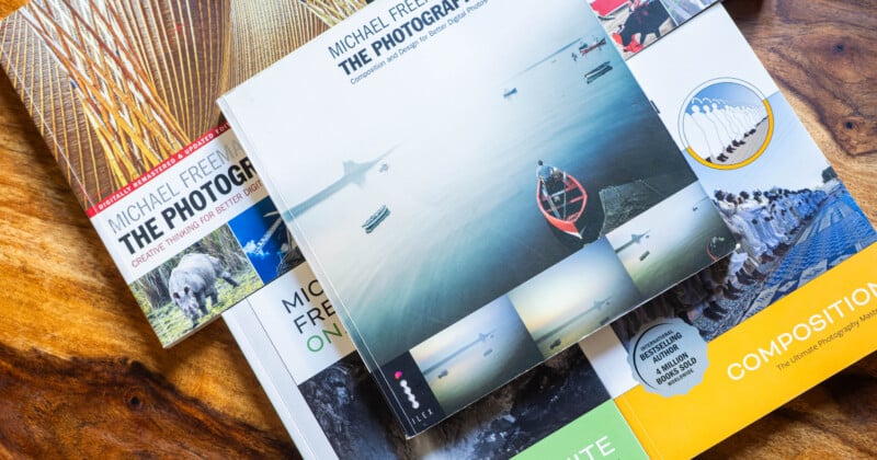 A collection of photography books by Michael Freeman are spread out on a wooden surface, with colorful covers featuring images of landscapes, animals, and people. The central book displays a serene water scene with a red boat.