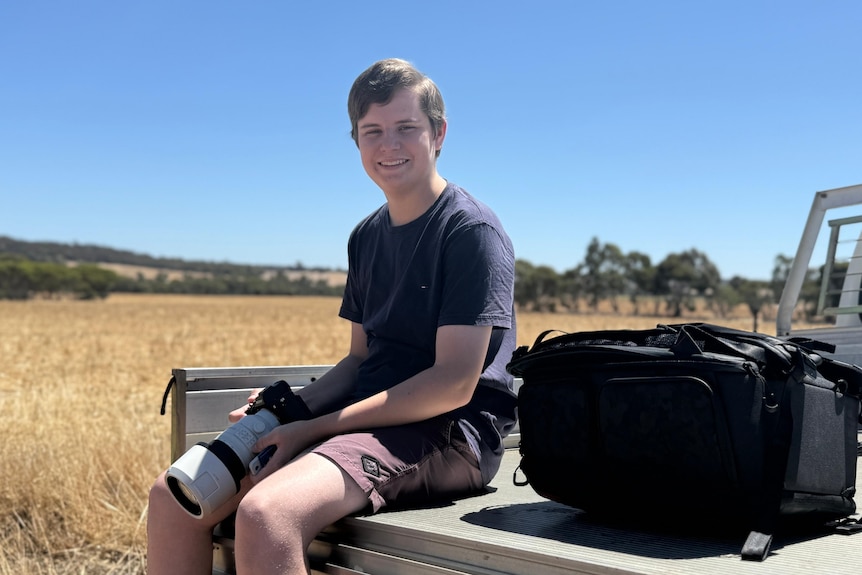 A smiling young man holds a camera with a telescopic lens as he sits on the back of a ute on a country property.