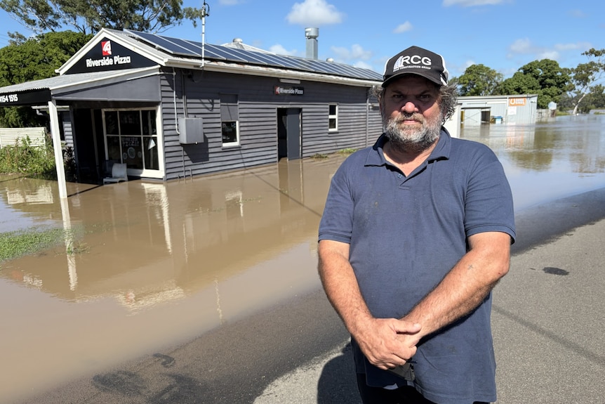 bearded man in navy polo frowning at camera in front of flooded grey building in brown water