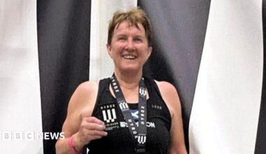 Carole Munro smiles at the camera wearing a medal and holding a patch. She is standing in front of a black and white striped backdrop and is wearing a black vest top.