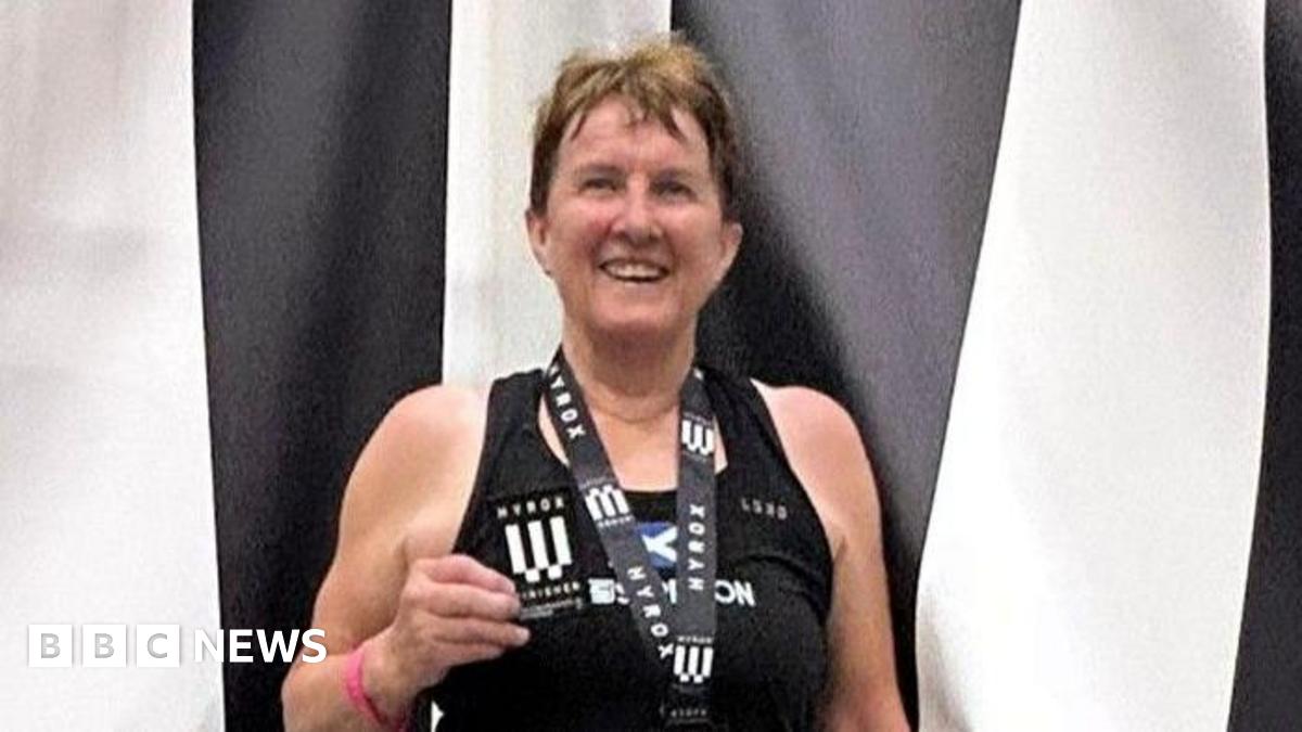 Carole Munro smiles at the camera wearing a medal and holding a patch. She is standing in front of a black and white striped backdrop and is wearing a black vest top.