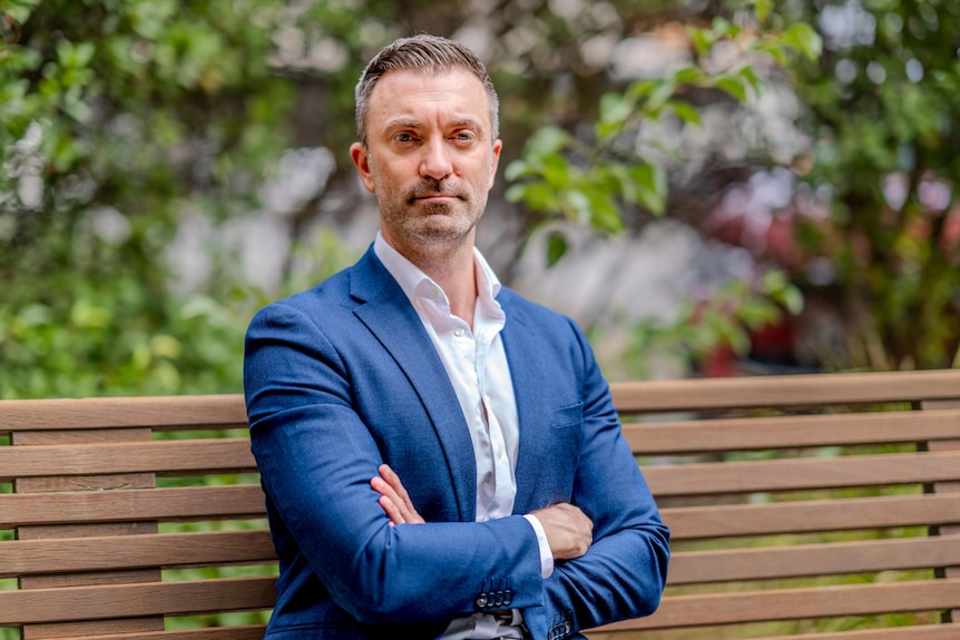 A man sits with his arms folded and a serious expression on a wooden bench with trees in the background